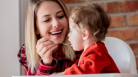 young mother feeding a child with porridge. Toddler baby sitting in armchair. Brick wall on...