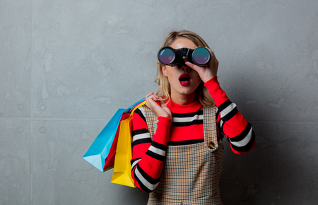 Portrait of a young style blonde girl in red striped sweater with binoculars and shopping bags on grey background.の写真素材