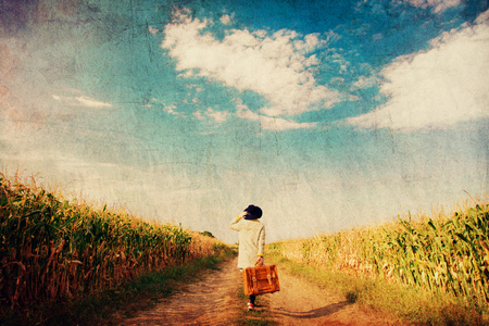 Young girl coat with suitcase on a rural road near cornfield. Image in old dirty paper styleの写真素材