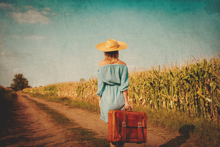 Young girl in dress and hat with suitcase walking along a rural road. Image in old dirty paper styleの写真素材