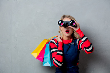 Portrait of a young style blonde girl in red striped sweater with shopping bags and binoculars on grey background.の写真素材
