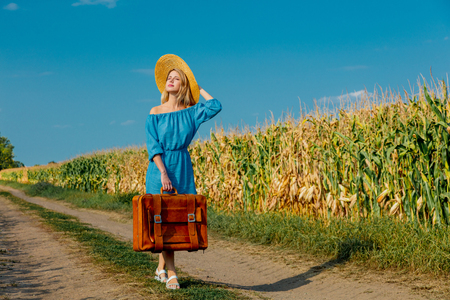 Young girl in dress and hat with suitcase walking along a rural road.の写真素材