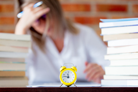 Tired student girl in shirt with books and alarm clock on foregroundの写真素材