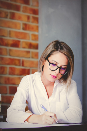 Young businesswoman in glasses sitting on working place with pen. Brick wall on backgroundの写真素材
