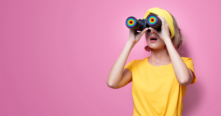 Young redhead girl in yellow t-shirt and blue jeans holding binoculars on pink background