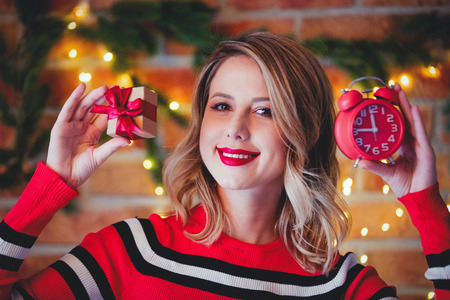 Portrait of a young girl in red striped sweater with gift box and alarm clock at Christmas lights...