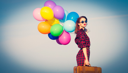 Beautiful girl in plaid dress with multicolored balloons and bag on blue sky backgroundの写真素材