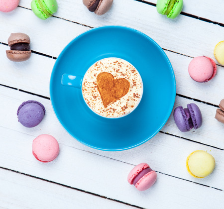 Cup of Cappuccino with heart shape symbol and macaron on white wooden backgroundの写真素材