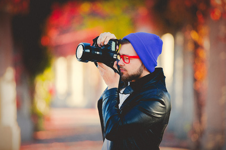 Young white guy in glasses and hat make a shoots with a camera in a park with yellow trees on...