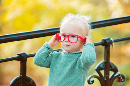 little white blonde girl in red glasses and green sweater in a park. Autumn seasin timeの写真素材
