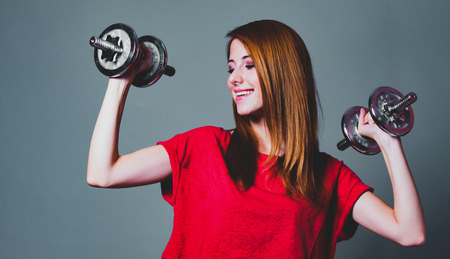 Sport woman in red shirt with dumbbells on gray background