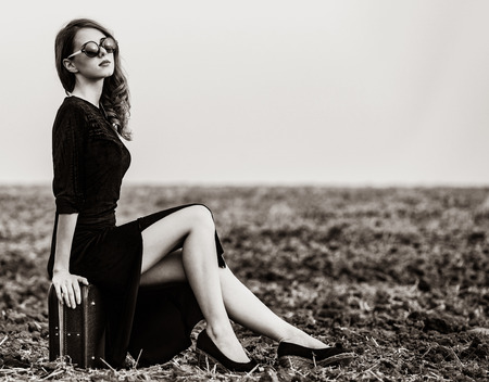 Fashion woman in black dress and sunglasses sitting on suitcase at field after harvest. Image in...