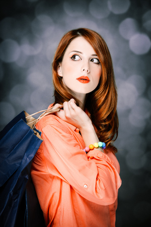 Beautiful surprised caucasian redhead adult girl in coral shirt and shopping bags on gray backgroud.の写真素材