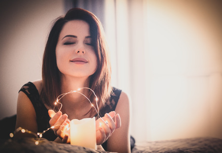 Portrait of beautiful young woman holding garland and candle and lying on the bed on the wonderful window and curtains backgroundの写真素材