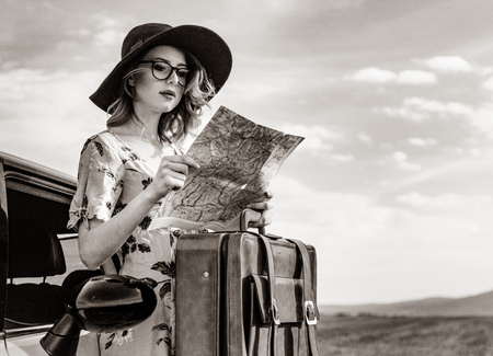 Portrait of young woman in dress with map and suitcase near a car at autumn countryside . Image in...