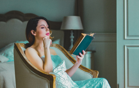 Young redhead woman sitting in chair in bedroom and reading book.の写真素材