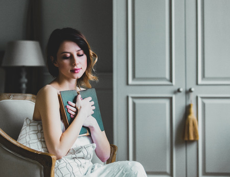 Young redhead woman sitting in chair in bedroom and reading book.の写真素材