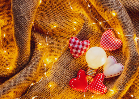 Heart shape gifts and bulb with Fairy Lights around on jute background. Above point of view