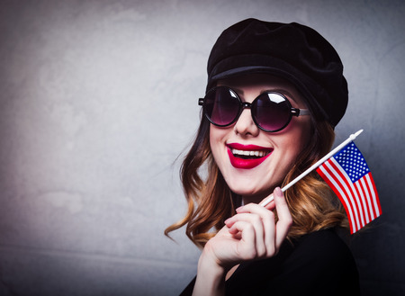 Style redhead girl in black hat and clothes in sunglasses with flag of the United States of America on grey background.の写真素材