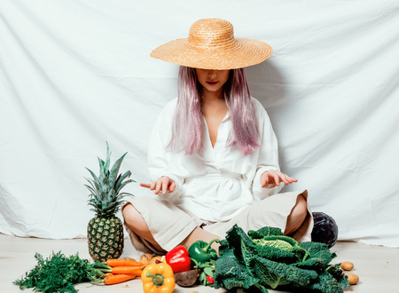 Beautiful caucasian vegan woman in hat with vegetables sitting on a flooor. Spring time concept and healthy mealの写真素材