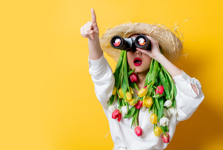 Beautiful woman in white shirt and hat with fresh springtime tulips instead hair and bonocular on...