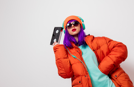 Portrait of a beautiful girl with purple hair in orange hat and jacket and with headphones and VHS cassette on white background. Trendy styleの写真素材