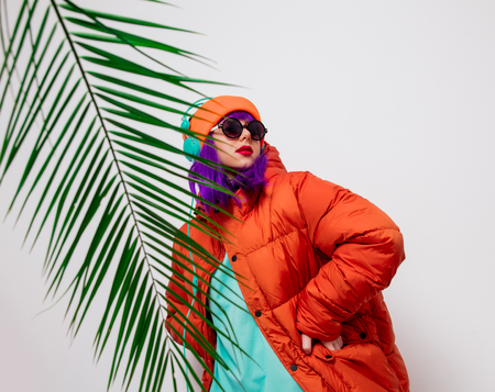 Portrait of a beautiful girl with purple hair in orange hat and jacket and with headphones on white background, palm branch. Trendy styleの写真素材