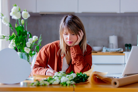 Businesswoman in modern clothes writing something in a note while wrapping a white roses and paper as a bouquet for Holiday. St. Valentines Day or springtime conceptの写真素材