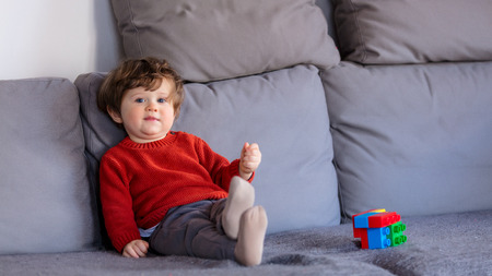 Funny little boy sitting on sofa in red sweater. Side view