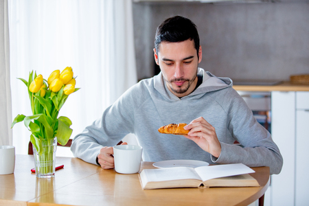 Young handsome man sitting at table with cup of coffee or tea and eating  croissant. Kitchen location.の写真素材