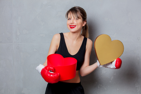 Young woman in boxing gloves with heart shape gift box in hands on gray backgroundの写真素材