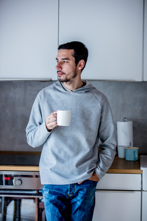 Young man with cup of coffee staying at kitchen at home. Candid portraitの写真素材
