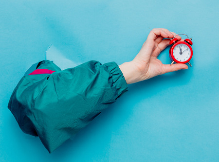 Female's hand in 90's style jacket holding alarm clock on blue background