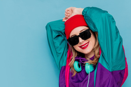 Portrait of a woman in red hat, sunglasses and suit of 90s with headphones on blue background.