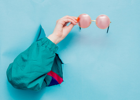 Female's hand in 90's style jacket holding sunglasses on blue backgroundの写真素材