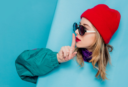 Portrait of a woman in red hat, sunglasses and suit of 90s on blue background.