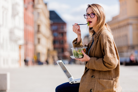 Young businesswoman eating salad and working with computer at urban outdoor. Dinner time conceptの写真素材