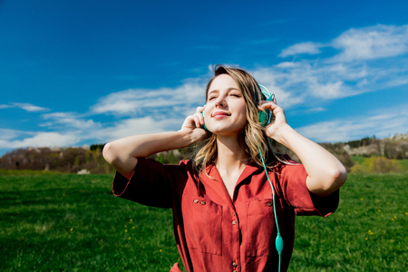 Beautiful girl in red dress and hat have a carefree time with headphones on meadow in mountains with forest on background. Spring seasonの写真素材