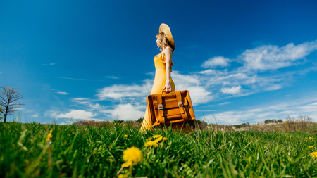 Beautiful girl in yellow dress and suitcase on mountain meadow with dandelions. Spring seasonの写真素材
