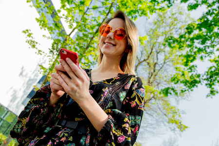 Young woman in sunglasses with mobile phone sitting on green alley near modern building of Universityの写真素材