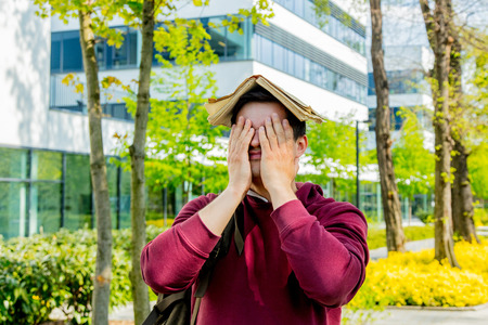 Young upset student with book on head stay near modern building of University. Spring time seasonの写真素材