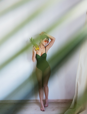 Female in bikini holding pineapple and with palm leaves out of focus on grey backgroundの写真素材