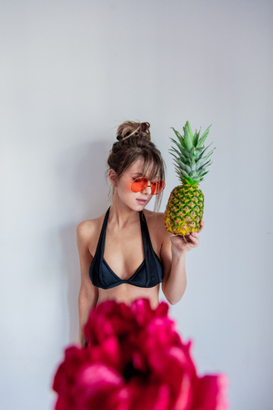 Adult woman in bikini with pineapple and sunglases. Peony flower on foreground.の写真素材
