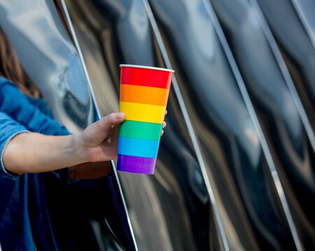 female hand holding a cup in the color of the LGBT flag on the background of metal construction
