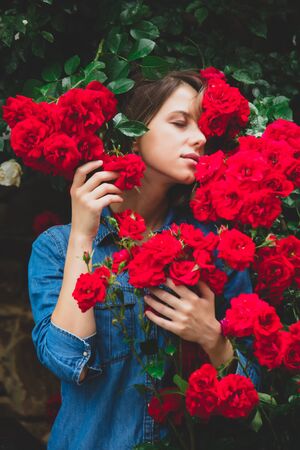 Young woman near the bush of red roses in a garden. Side viewの写真素材