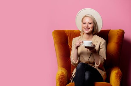Beautiful blonde woman in cowboy hat sitting in a vintage armchair with cup of coffee on pink backgroundの写真素材