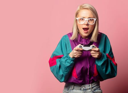 Beautiful blonde woman in 90s clothes with joystick on pink background