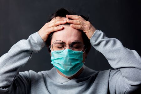 young white man in glasses and protective mask with a headache on dark background