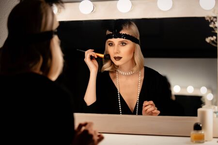 Beautiful blonde woman in twenties years clothes applying makeup near a mirror with bulbs 