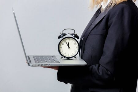 Businesswoman in black suit and face mask with alarm clock and computer on grey backgroundの写真素材